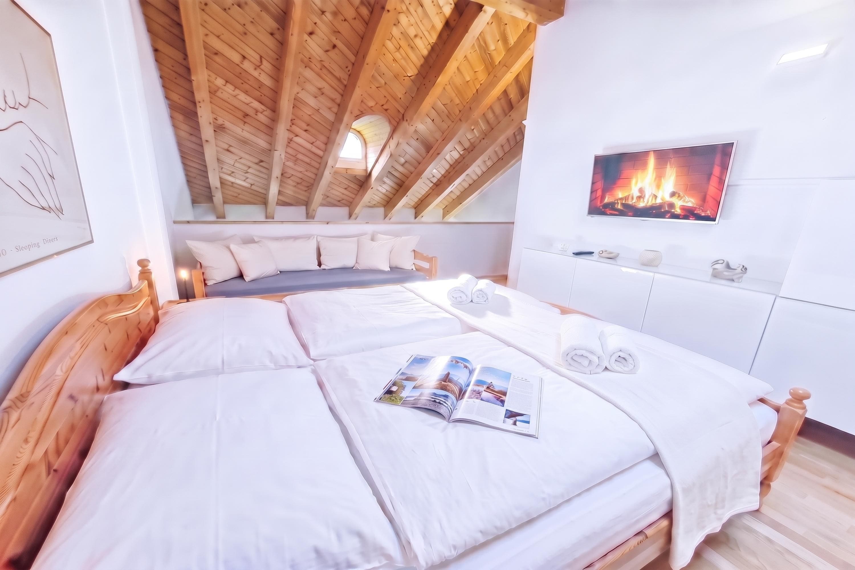 Ein Schlafzimmer mit schrägem Dach, einem Bett mit weißer Bettwäsche, einer Zeitschrift auf dem Bett, einem Kamin an der Wand und einem Fenster mit Aussicht.
