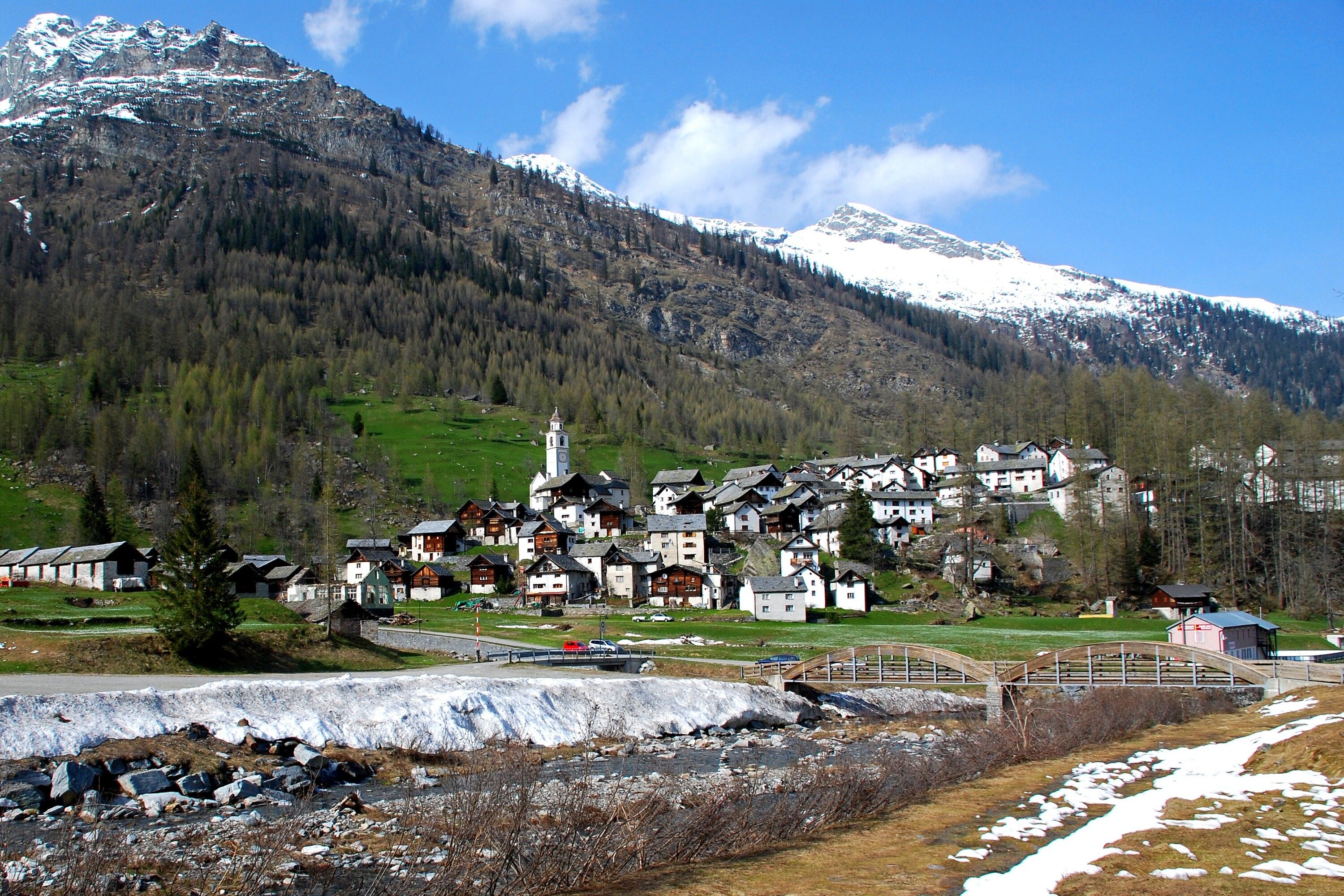 Un village de montagne aux maisons traditionnelles, avec un pont et des sommets enneigés en arrière-plan.