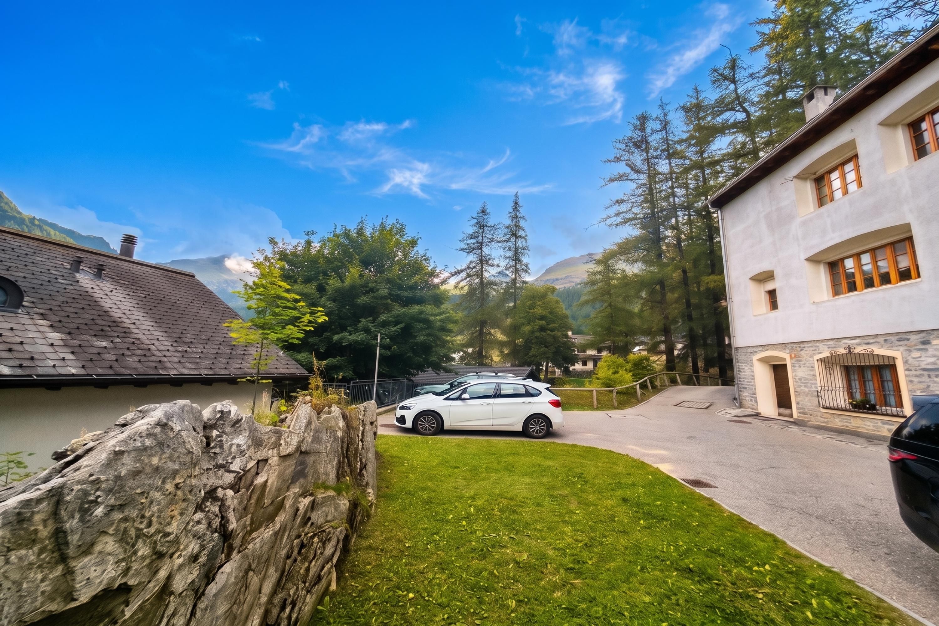 A residential area with a white car parked on a paved driveway, surrounded by lush greenery and a stone wall.