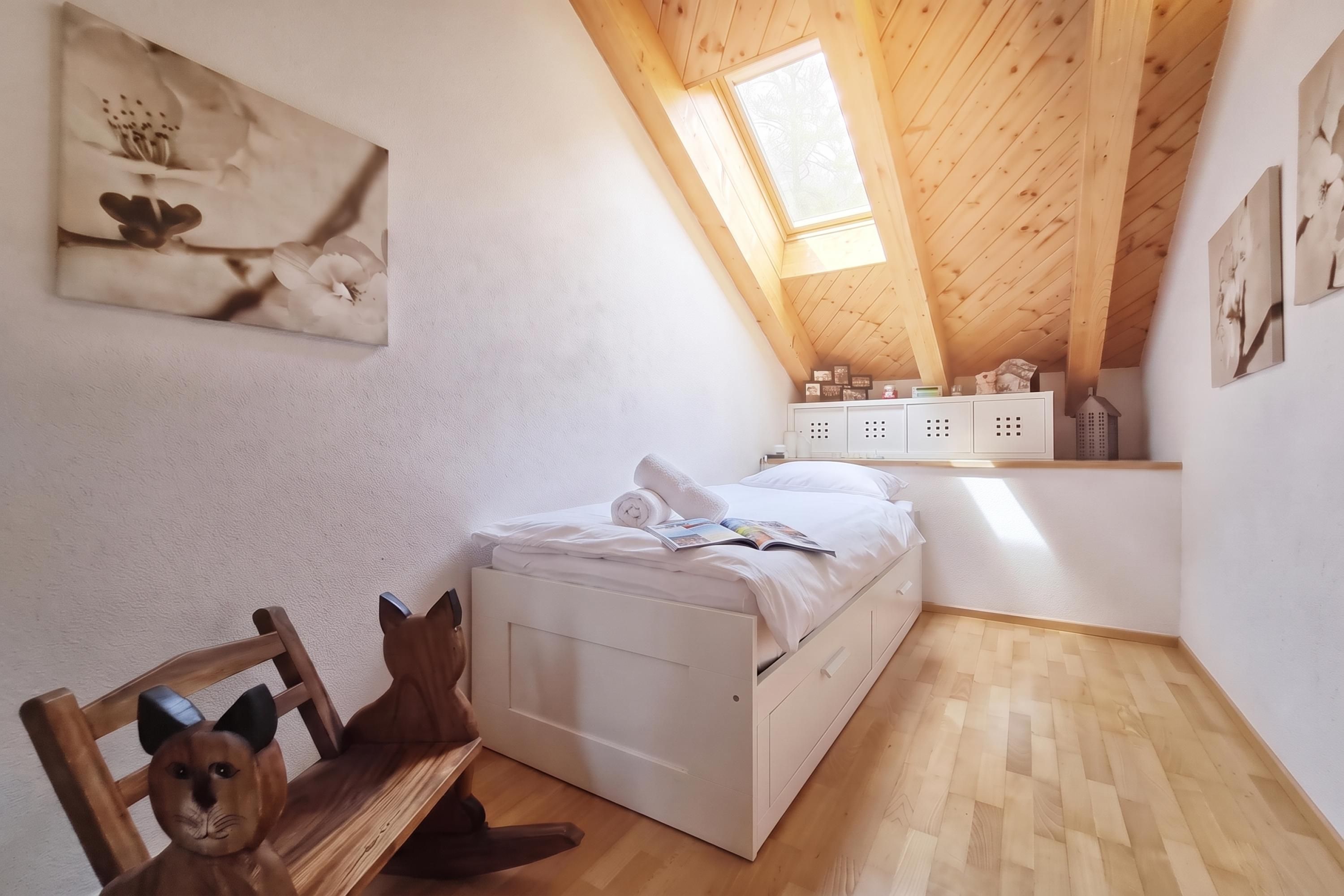 Ein Schlafzimmer mit schrägem Dach, einem Bett, einem Stuhl und einem kleinen Regal mit Gegenständen.