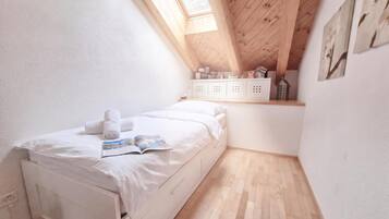Una stanza piccola e luminosa, con soffitto in legno, un letto con biancheria bianca, un piccolo ripiano con diversi oggetti e una finestra che fa entrare luce naturale.