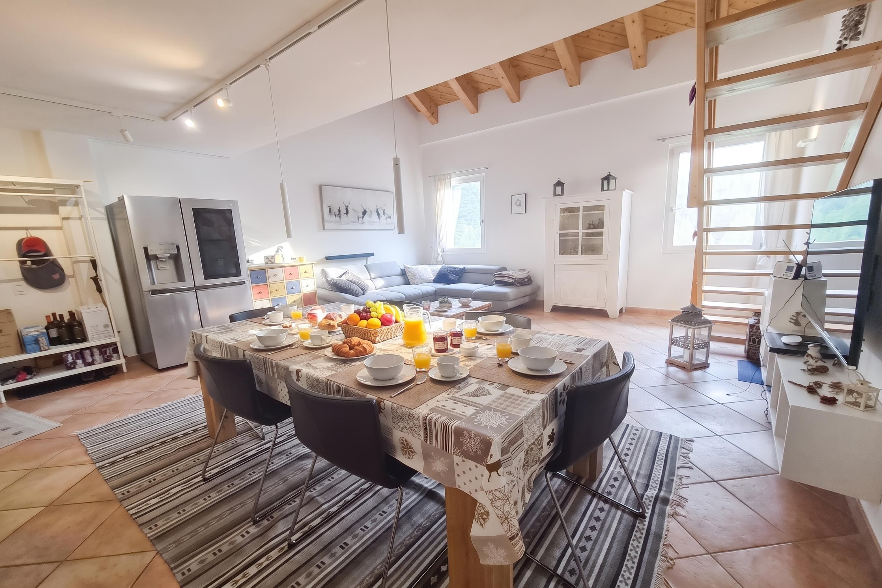 Ampla sala de estar com mesa de jantar posta para o café da manhã, um sofá e uma cozinha com geladeira.