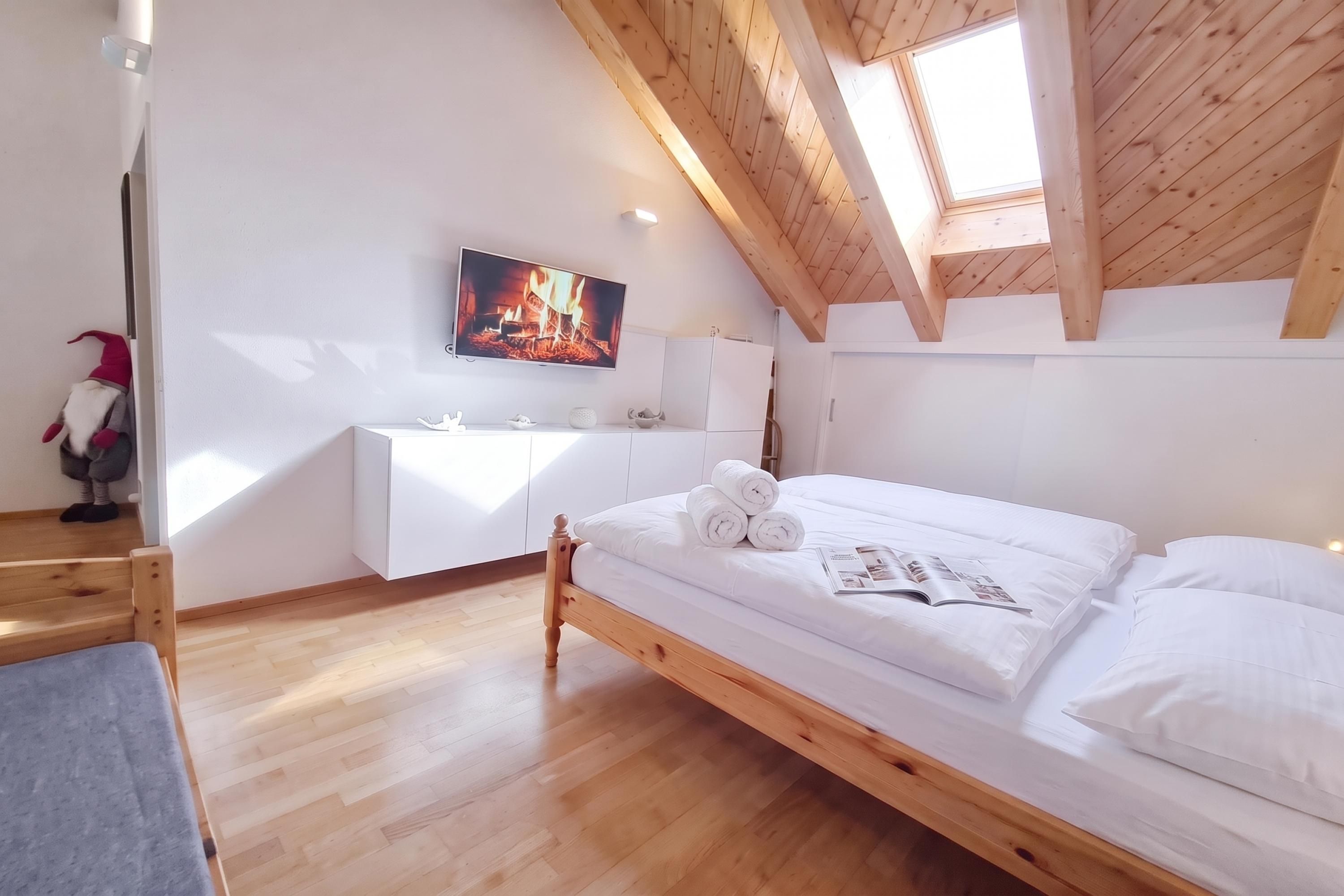Une chambre avec un plafond en bois, un lit recouvert de draps blancs, un téléviseur à écran plat et une petite commode blanche.