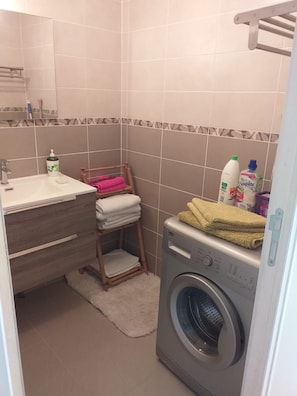 Bany compacte amb rentadora, un tamboret de fusta amb tovalloles, un lavabo amb productes de tocador i un respatller de rajola.
