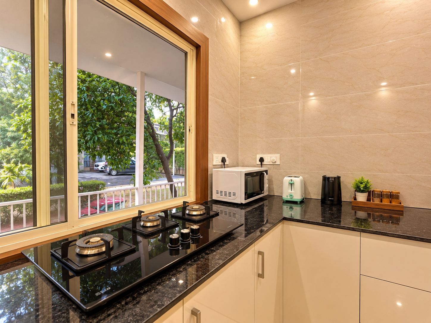 A modern kitchen with a gas stove, microwave, toaster, and a view of a street with trees and parked cars.