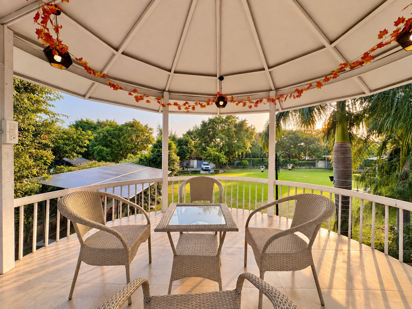 Un espacio de estar al aire libre cubierto, con mobiliario de mimbre y una mesa de vidrio, con vistas a un jardín y paneles solares.