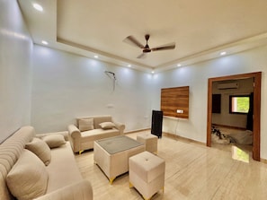 A modern living room with a sofa, ottoman, and coffee table, a ceiling fan, recessed lighting, and a mounted TV. - Mulberry Farm (Noida)