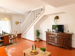 Ein Wohnzimmer mit einer Holztreppe, einem Essbereich, einem Fernseher auf einer Holzkommode und einem kleinen Tisch mit Flaschen und Pflanzen.