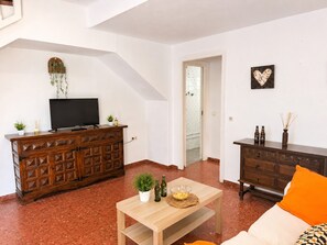 Ein Wohnzimmer mit Fernseher auf einer Holzkommode, einem kleinen Tisch mit Schüssel und Flaschen sowie einem Sofa mit orangefarbenem Kissen.
