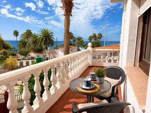 A balcony with a table and chairs, overlooking a coastal area with palm trees and a clear sky. - Villa Juanita en Nerja con Vistas al mar (Nerja)