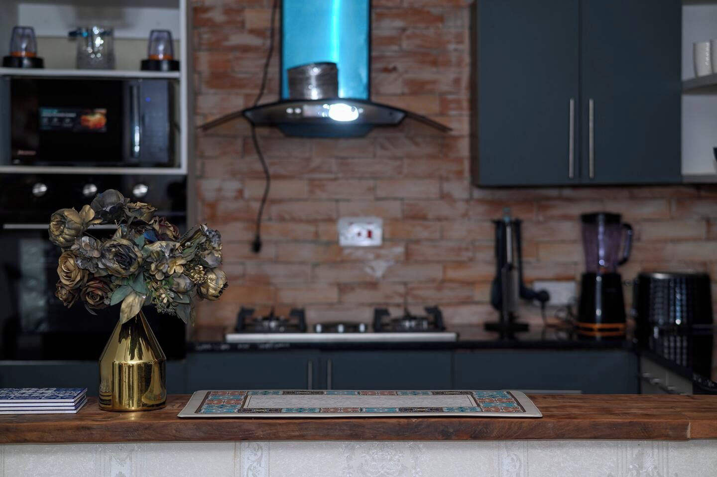 Una cocina con pared de ladrillo, estufa a gas, cafetera y un florero con flores secas en la encimera.
