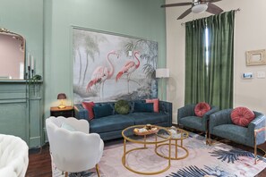 Ein Wohnzimmer mit einem blauen Sofa, zwei Sesseln, einem weißen Stuhl, einem runden Couchtisch, einem Flamingo-Gemälde, einem Deckenventilator und einem grünen Vorhang.