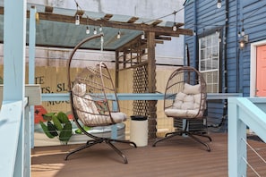 Two wicker chairs with cushions on a wooden deck, a white trash bin, and a blue building in the background. - Sleeps 6 // Backyard+Cowboy Pool+Firepit+Mural+ NEAR FQR At Flamingo Haus (New Orleans)