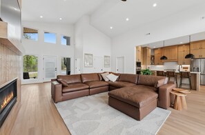 A modern living room with a sectional sofa, a fireplace, and a view of the kitchen. - Trailside MTB Retreat with Sauna (Bella Vista)