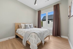 Ein Schlafzimmer mit einem Holzbett, einem Fenster mit Vorhängen und Blick auf ein Haus.