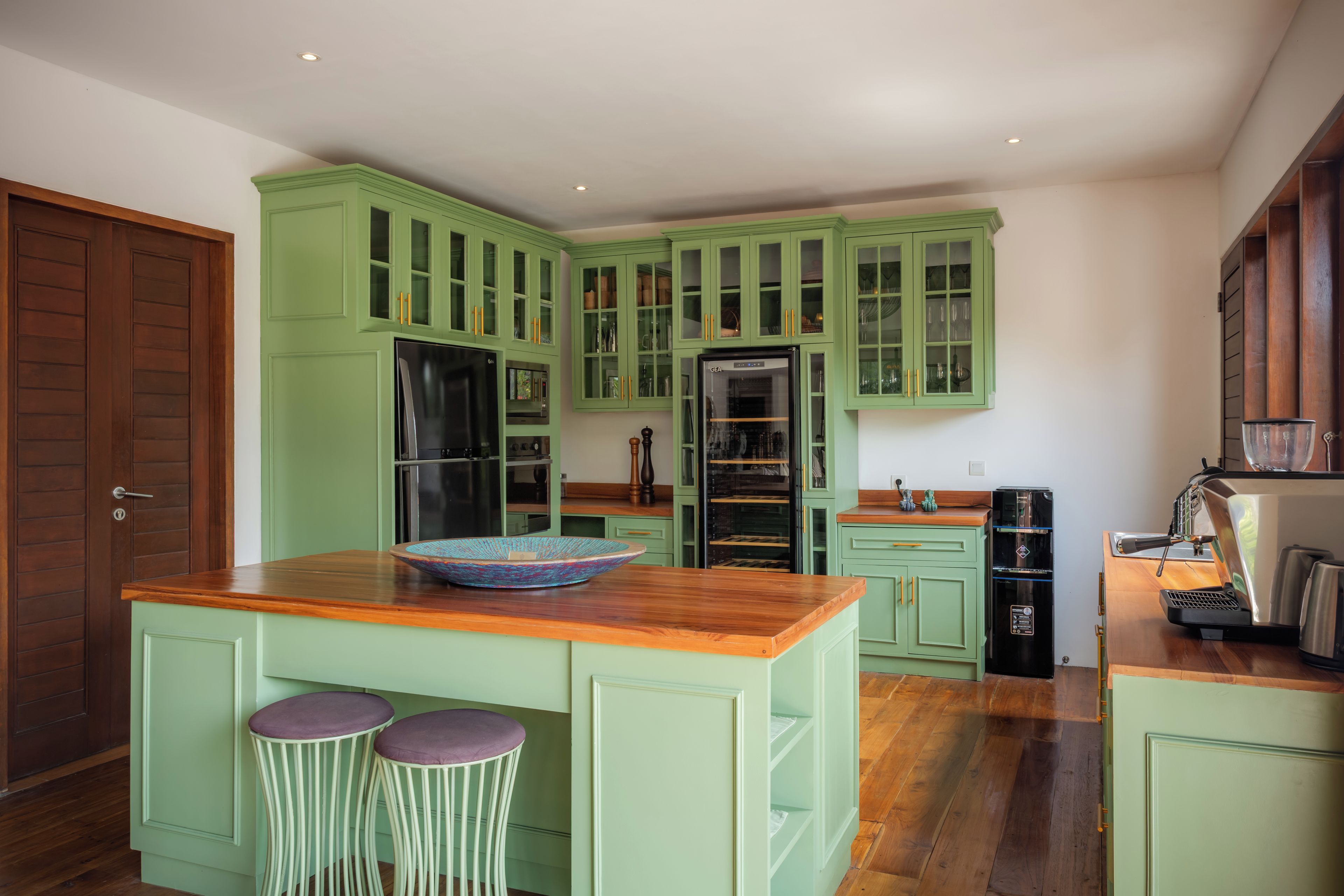 Una cocina con armarios verdes, un refrigerador negro, una máquina de café e isla de madera con dos taburetes.