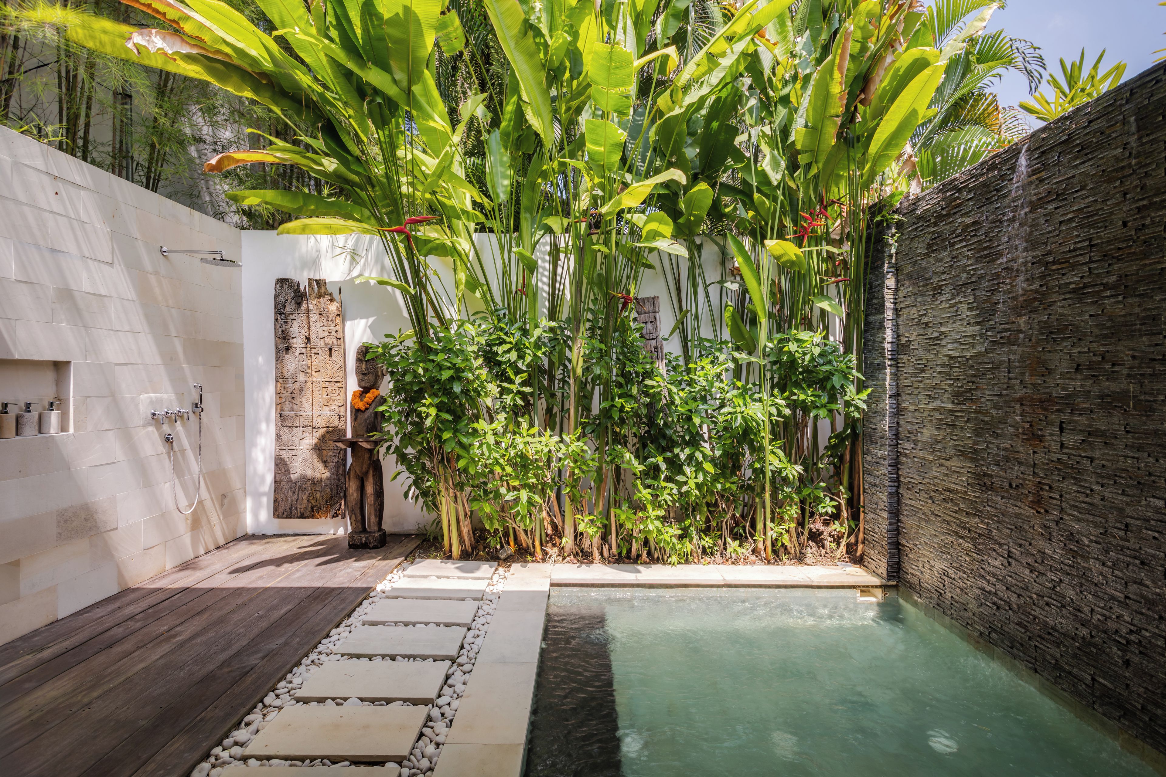 Zona privada de piscina con un sendero de piedra que lleva a una piscina pequeña, rodeada de exuberante vegetación tropical y un muro blanco con un panel decorativo.