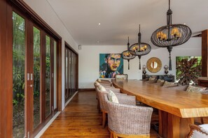 A spacious dining area with a long wooden table and wicker chairs, large windows, and a modern chandelier. - Villa Naty Luxury 6 Bedrooms Near Canggu (Bali)