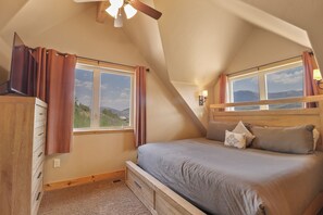 A bedroom with a sloped ceiling, a bed with a headboard, two windows with curtains, a ceiling fan, and a view of the outdoors. - 947 Blue Ridge 4 Bedroom Home (Silverthorne)