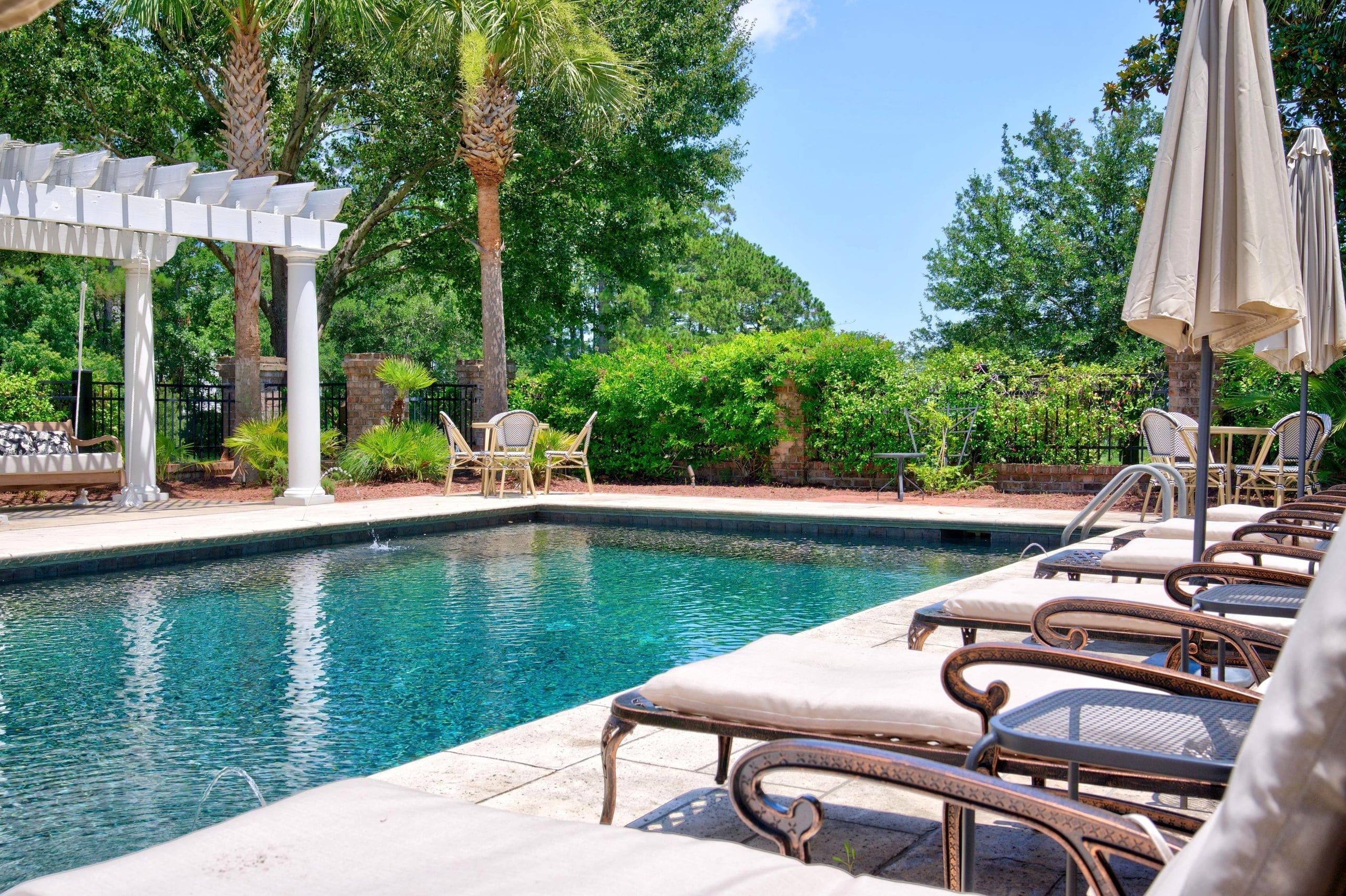 Un área de piscina con sillones de descanso y sombrillas, rodeada de árboles y una pérgola.