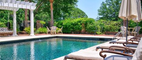 Un área de piscina con sillones de descanso y sombrillas, rodeada de árboles y una pérgola.