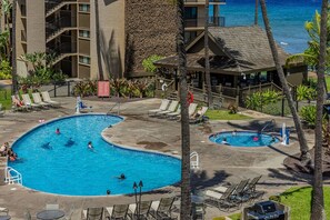 A hotel pool area with multiple swimming pools, lounge chairs, and a building with a stone facade. - Papakea C204 Condo (Lahaina)