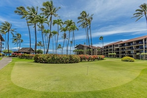 A golf course with a well-maintained lawn, palm trees, and a building with multiple balconies. - Papakea C204 Condo (Lahaina)