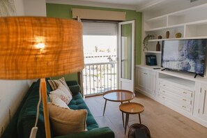 A living room with a green sofa, a TV, a small wooden table, and a balcony with a view. - The River Apartment with Balcony in the Heart of El Puerto de Santa María (El Puerto de Santa María)