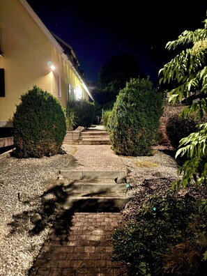 A nighttime pathway leading to a building with a yellow facade, flanked by neatly trimmed bushes and gravel.