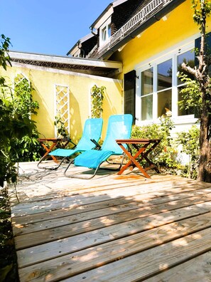 A wooden deck with two blue lounge chairs, a yellow building with a balcony, and a white window.