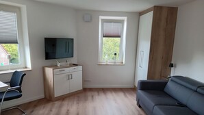 A modern living room with a flat-screen TV, a sofa, a desk, and a window with blinds. - Apartment in A Hotel 'Blickallee' (Havixbeck)