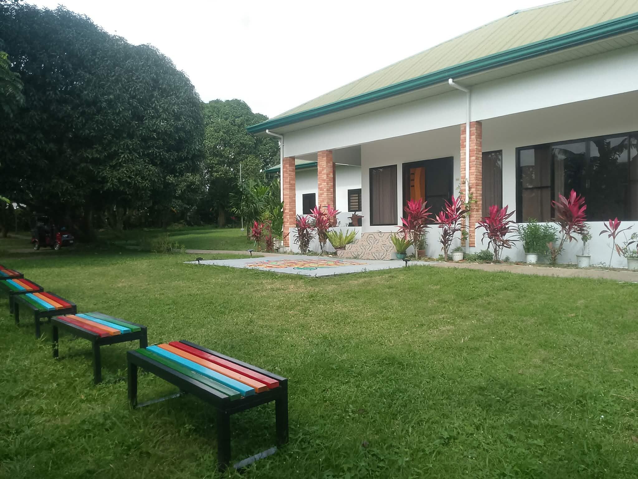 A building with a covered patio area, outdoor seating with benches, and a garden with various plants.