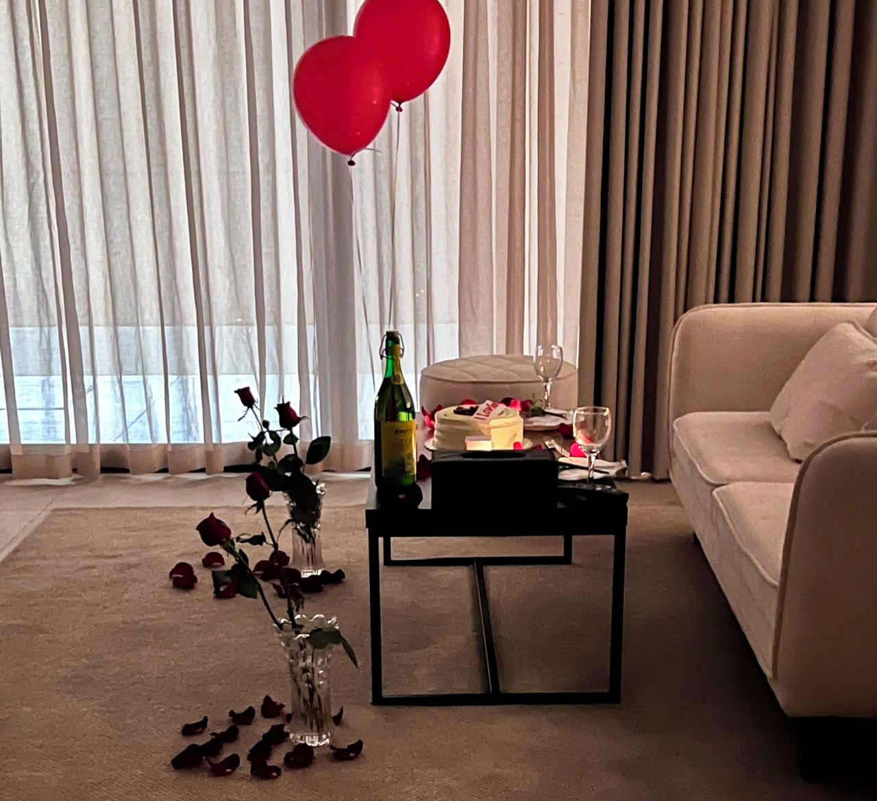 A living room with a sofa, a coffee table with a bottle and glasses, and a vase with flowers.