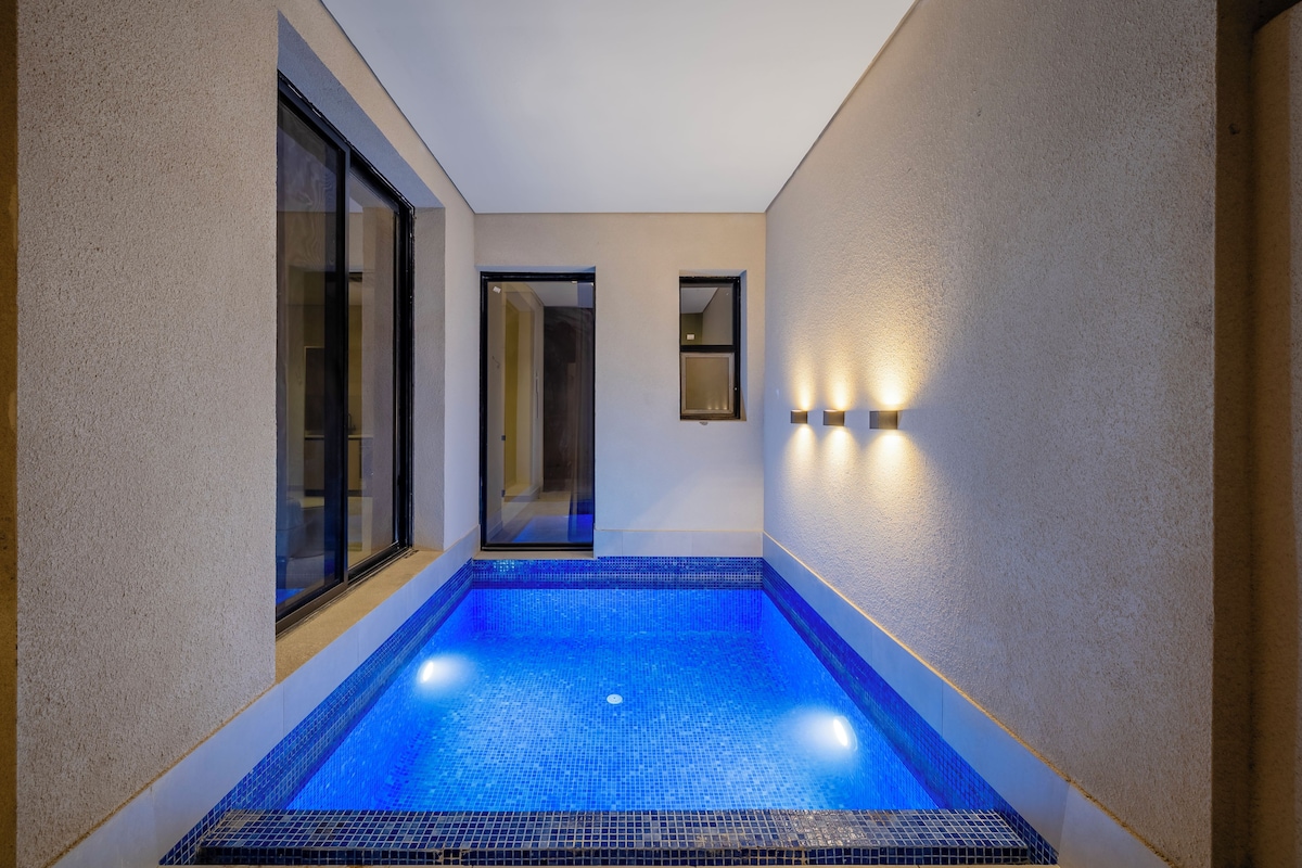 Una piscina interior estrecha con suelo y paredes de azulejos, una entrada con puerta de cristal y un pequeño espejo en la pared.