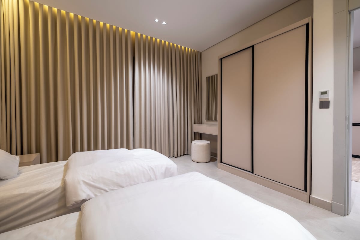 Una habitación de hotel moderna con una cama grande, una mesita de noche, un armario y un otoman redondo pequeño.