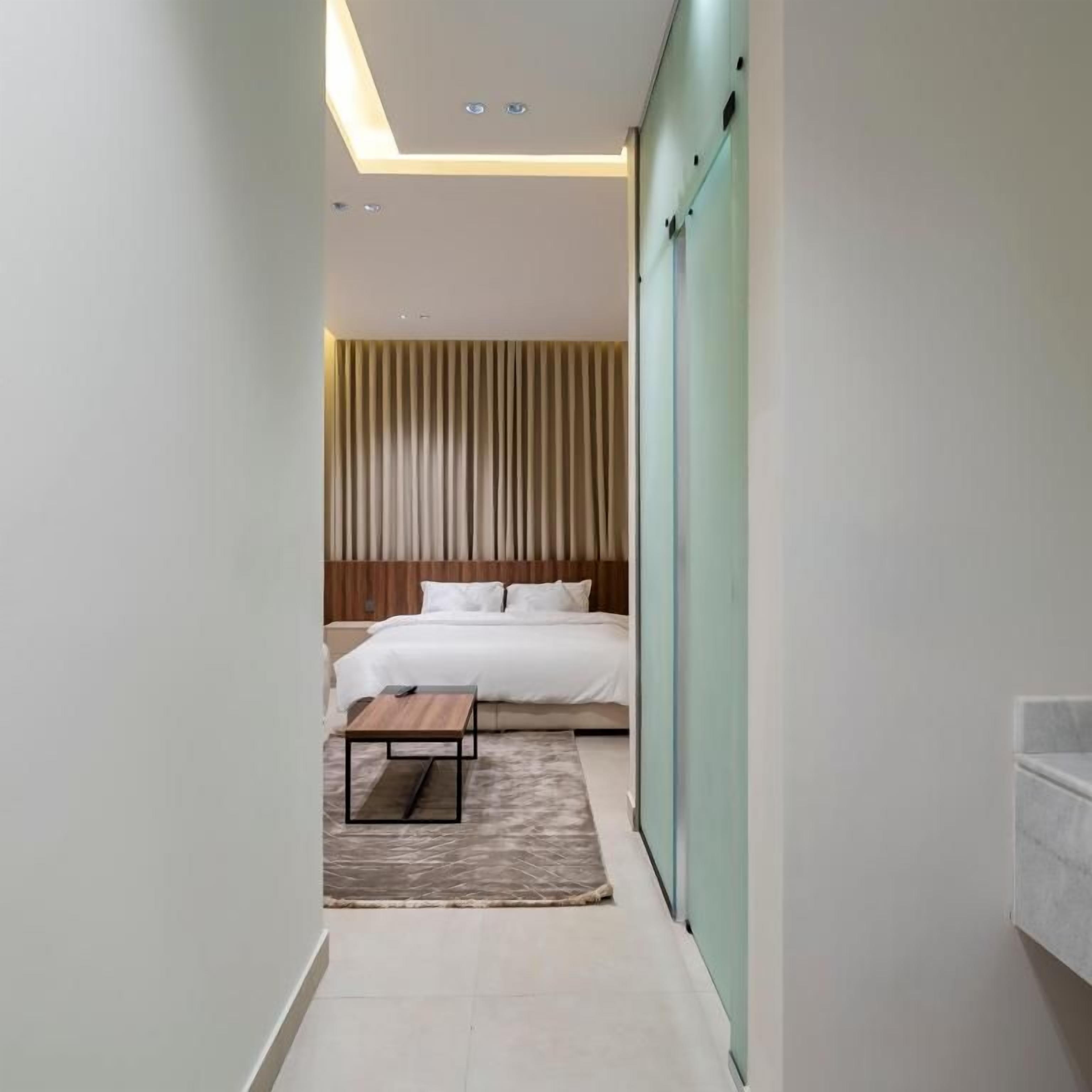 A narrow hallway leading to a bedroom with a bed, a wooden headboard, and a bedside table.