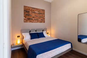 Een slaapkamer met een bed, nachtkastje, lamp, wanddecoratie en een spiegel.