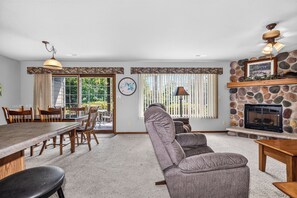 A living room with a fireplace, dining area, and a large window with blinds. - Eagle River Chain of Lakes Condo with Indoor Pool (Eagle River)