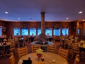 Ein Restaurant mit runden Tischen, Holzstühlen, einem zentralen Kamin und großen Fenstern.