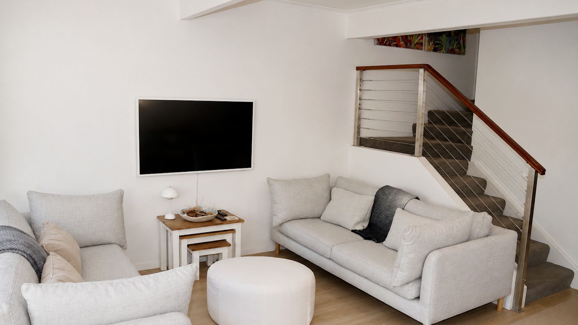 A modern living room with a sofa, ottoman, and a small side table with a lamp. A staircase with a wooden handrail leads to an upper level.