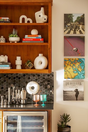 A kitchen with a wooden shelf holding books and decorative items, a hexagonal tile backsplash, and a framed photograph of a famous album cover. - Eastside Swim - Pool, Spa, Movie Room - Sleeps 13 (Austin)
