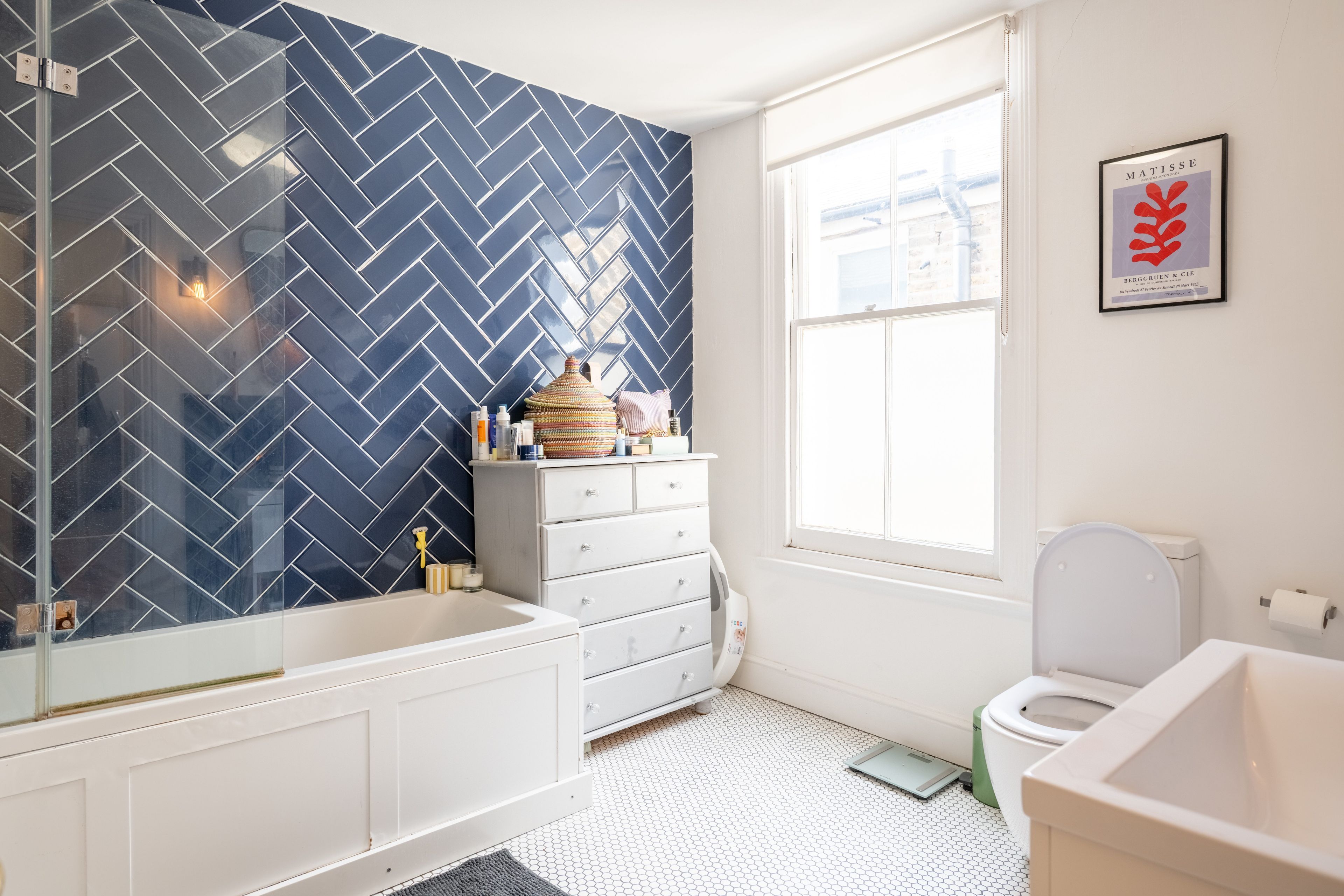 Une salle de bain avec un mur recouvert de carreaux disposés en motif chevron, un lavabo blanc, des toilettes blanches et une fenêtre donnant sur l’extérieur.