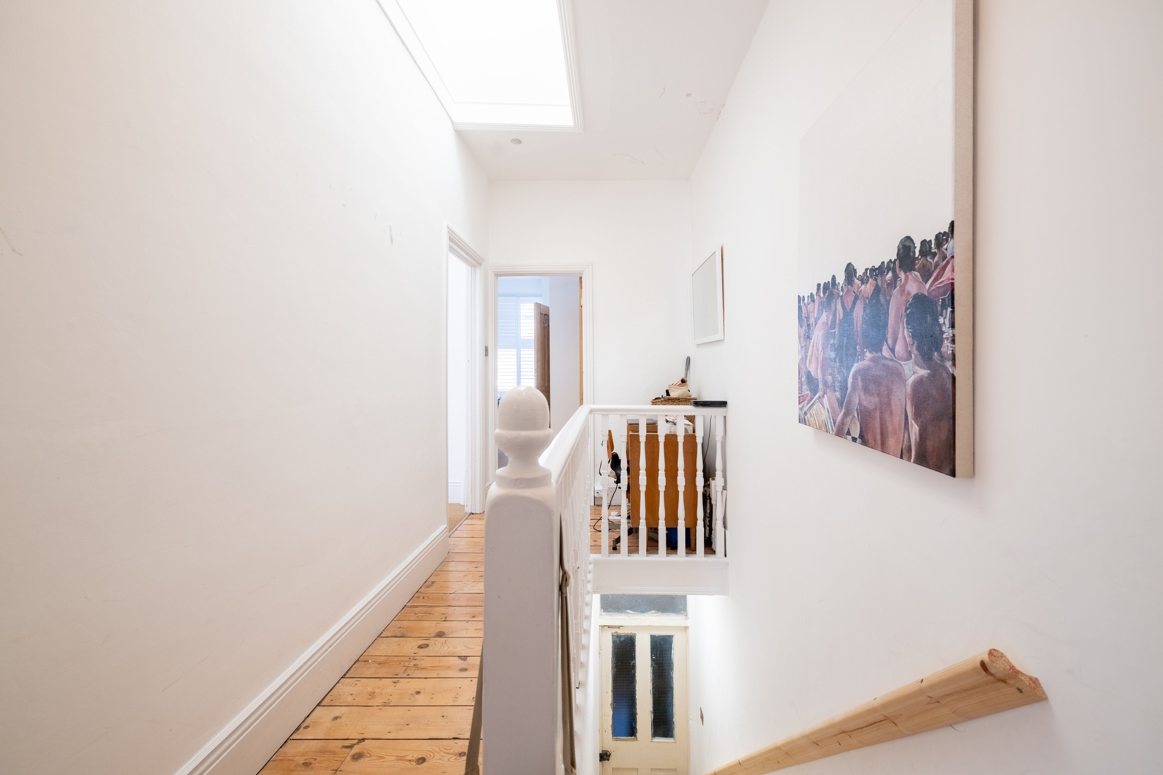 A narrow hallway with wooden flooring, white walls, and a white railing with a picture on the wall.