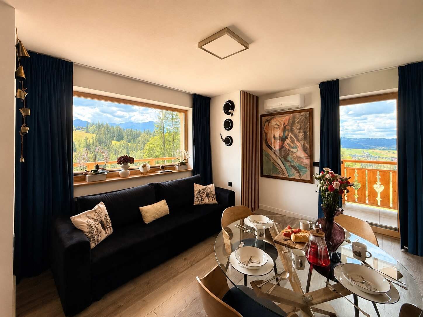 A modern living room with a black sofa, dining table, and chairs. There are large windows with a view of a forest and mountains.