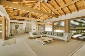 A spacious living area with a high ceiling and wooden beams, a sectional sofa, a coffee table, and large windows with blinds. - Kas Secreto – An Exclusive Oceanfront Escape Villa in Bonaire (Kralendijk)