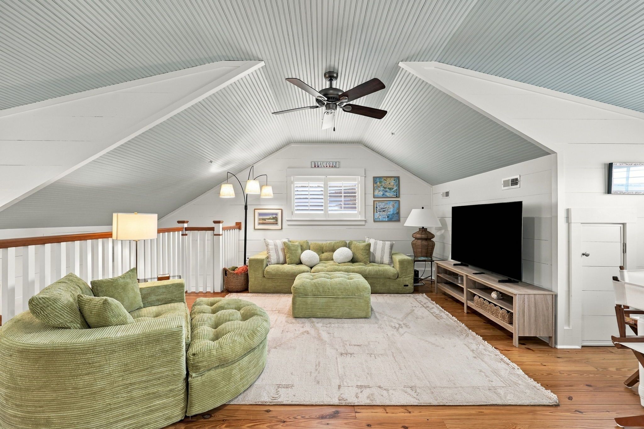 A spacious attic room with a sloped ceiling, a ceiling fan, a large flat-screen TV, a green sofa set, and a wooden floor.