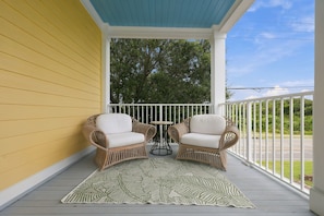 A covered porch with two wicker chairs and a small table, a patterned rug, and a view of the street and trees. - Charming Two-Level Coastal Condo near the Beaufort Hotel — Great Location! (Beaufort)