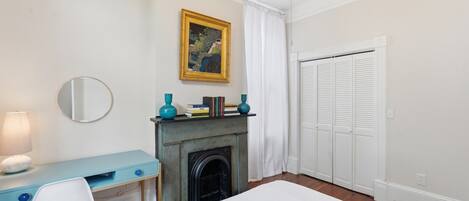 A bedroom with a bed, a desk with a chair, a fireplace, a wardrobe, and a ceiling fan.