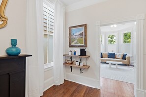A hallway with a wooden floor, a black fireplace, a blue vase, a painting, and a beige sofa in the adjacent room. - Charming Victorian 2BR Near Forsyth (Savannah)