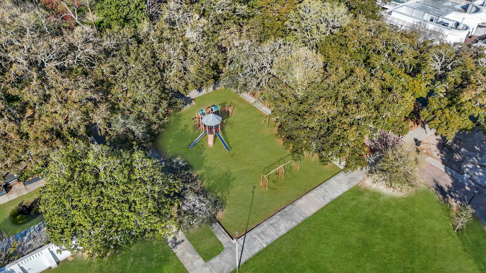Vue aérienne d’un parc avec une aire de jeux, entouré d’arbres et avec un bâtiment en arrière-plan.
